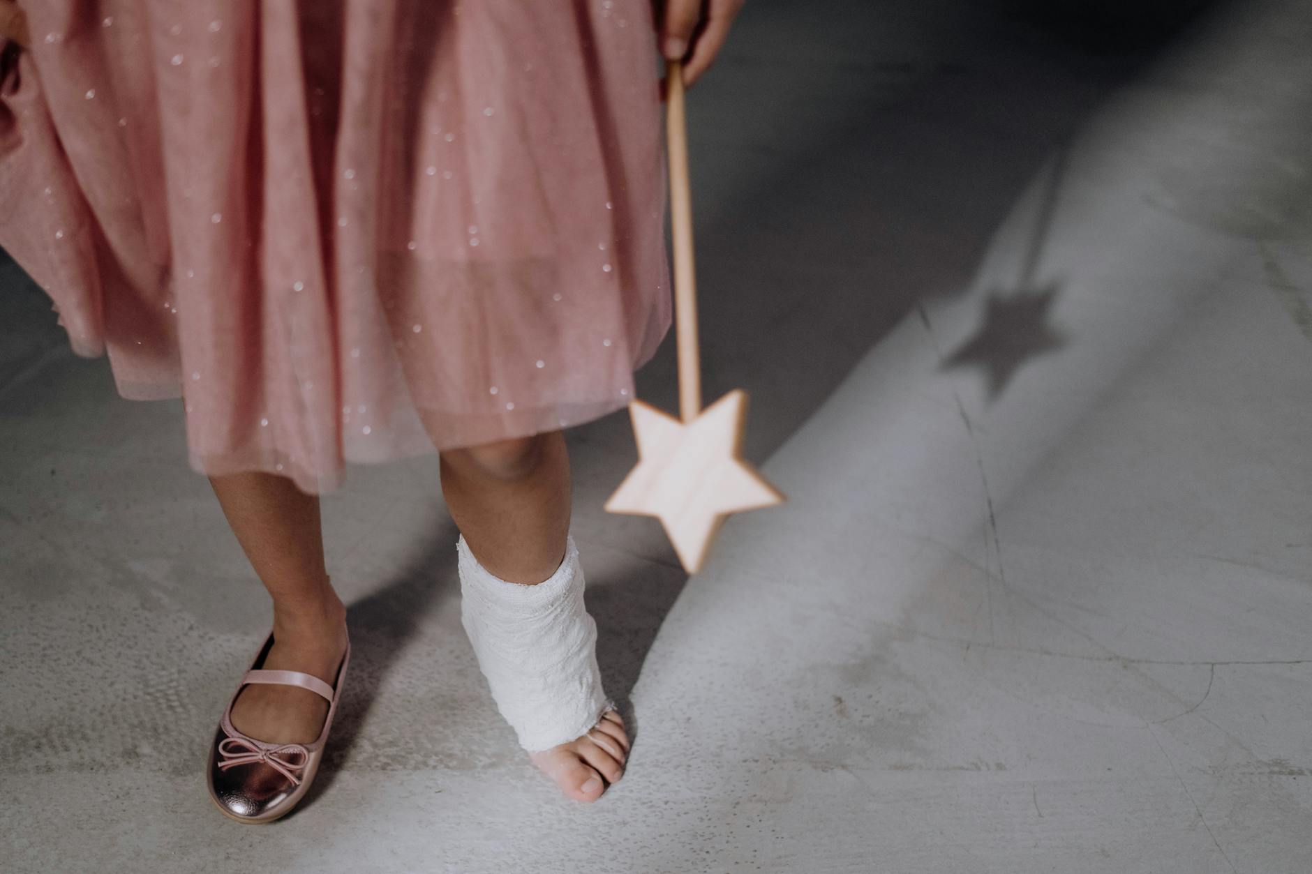 Dziewczynka w różowej sukience z nogą w gipsie trzyma drewnianą różdżkę