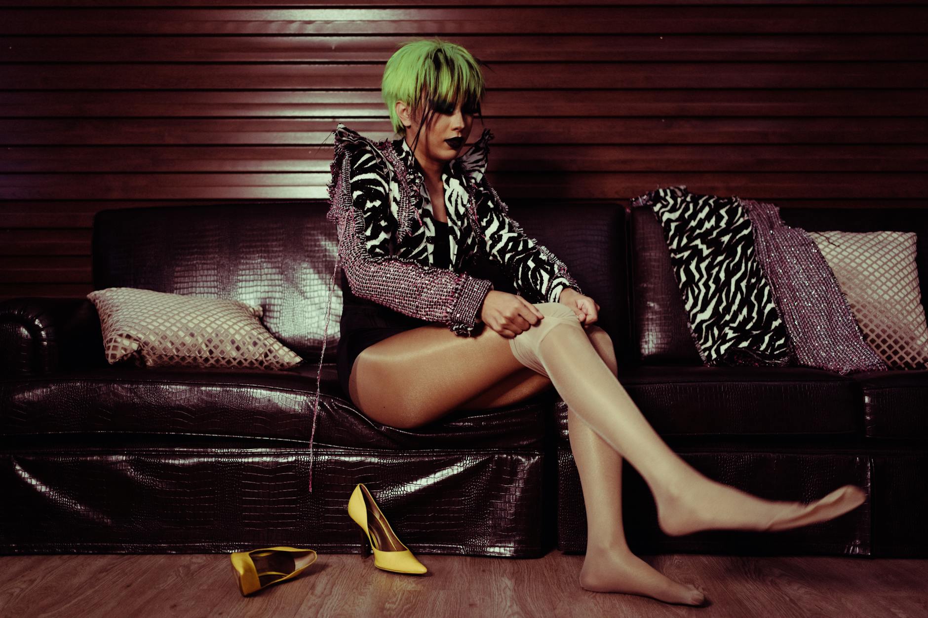 Kobieta w peruce zakłada rajstopy, siedząc na skórzanej sofie