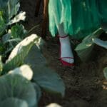 Osoba w zielonej sukience w czerwonych szpilkach stoi na polu kapusty